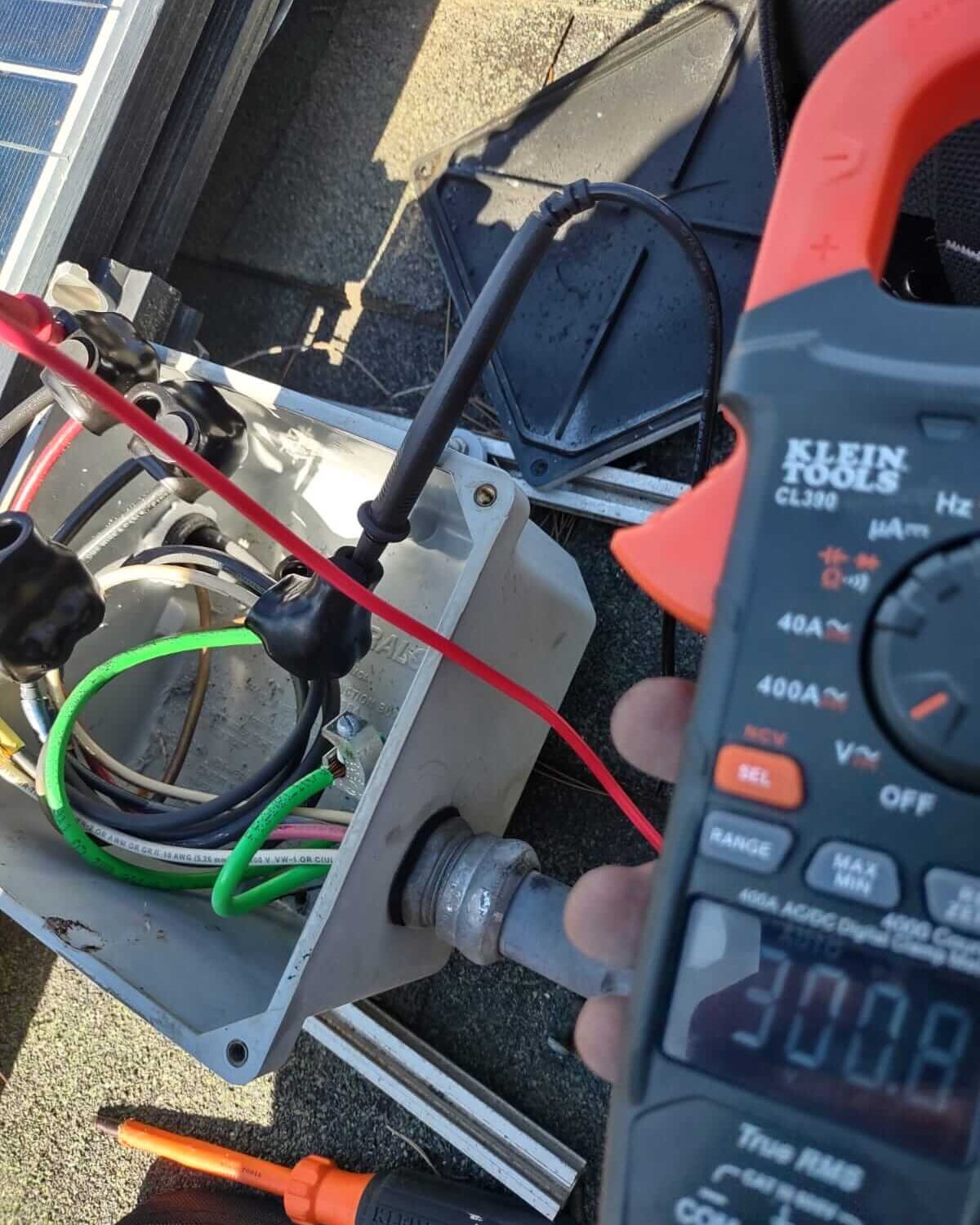 A clamp meter with test leads taking a live reading inside an opened rooftop junction box, beside the edge of a solar array on a composition shingle roof.